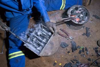 Man sat on floor in blue overalls soldering metal and surround by flakes of black carbon.